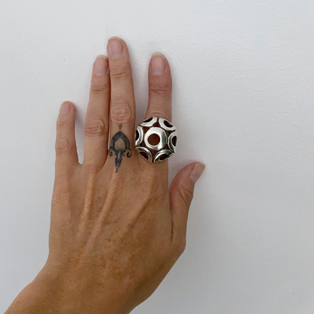 A hand wearing a large silver ring with a geometric, faceted pattern of black and white circular cutouts, set against a plain white background.