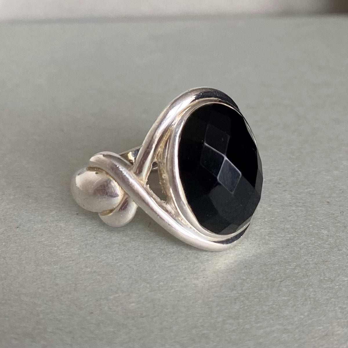 A silver ring featuring a large, faceted black onyx gemstone in a twisted wrap setting, photographed on a neutral gray surface.