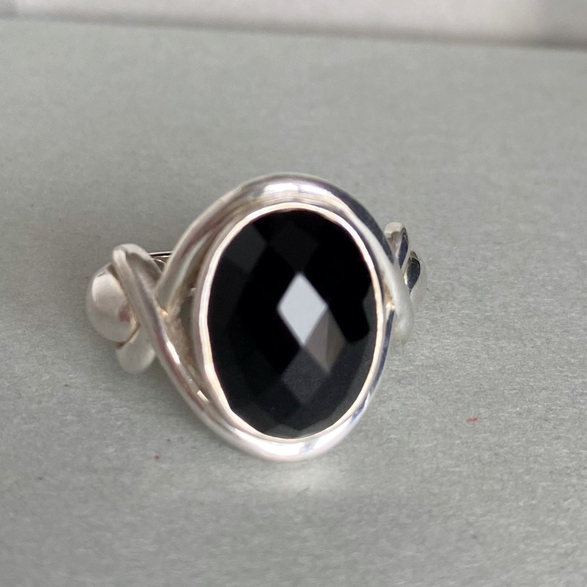 A silver ring featuring a large, faceted black oval gemstone, photographed on a neutral gray textured surface.