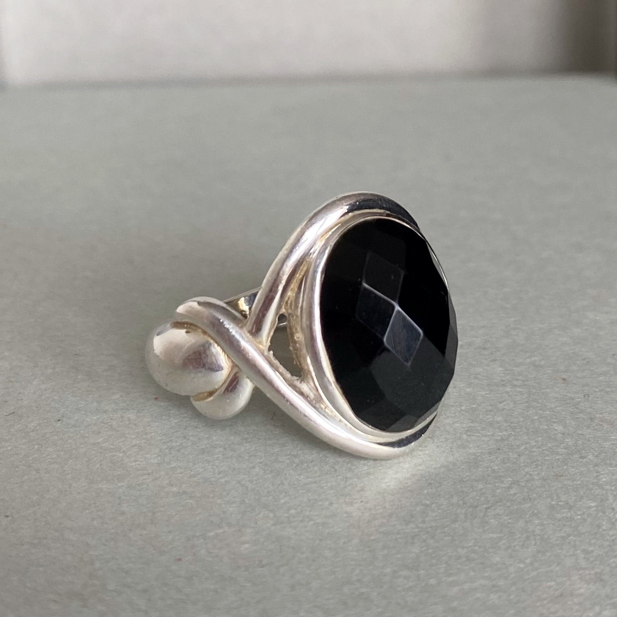 A silver ring featuring a large, faceted black gemstone in a wrap-style setting, resting on a neutral gray surface.