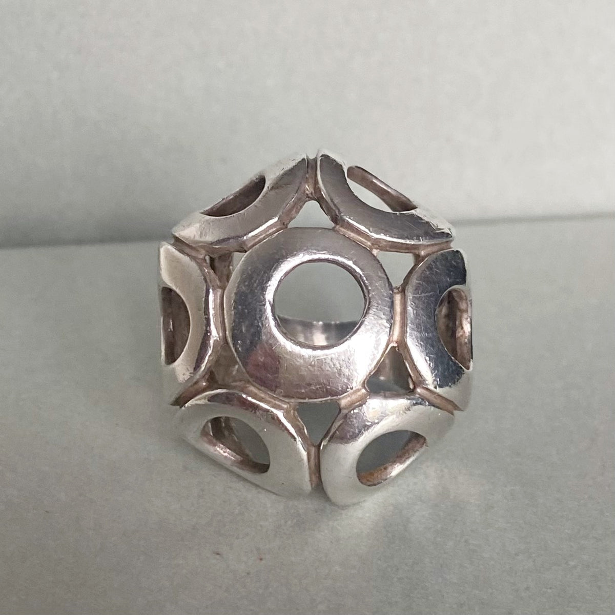 A silver geometric ring featuring a central circular cutout surrounded by interlocking petal-like segments with side cutouts, displayed on a neutral gray surface.