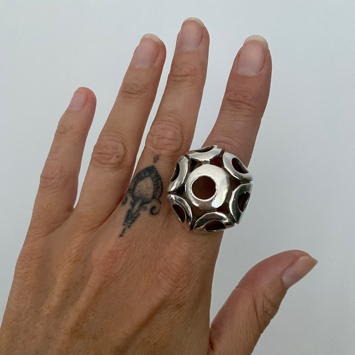 A silver geometric ring with circular cutouts worn on a finger, shown against a plain light gray background.