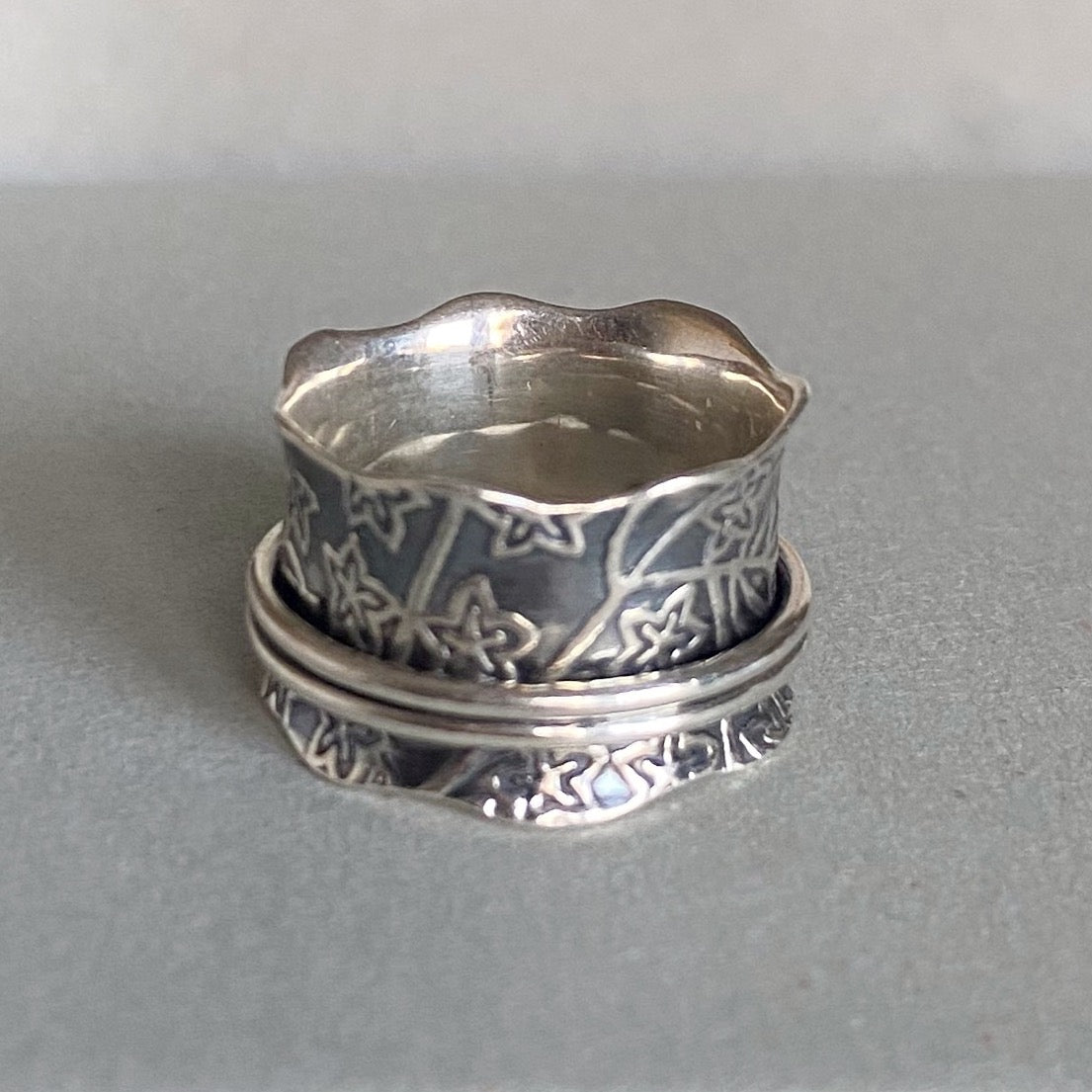 A sterling silver spinner ring with intricate floral and vine engravings, resting on a neutral gray surface.