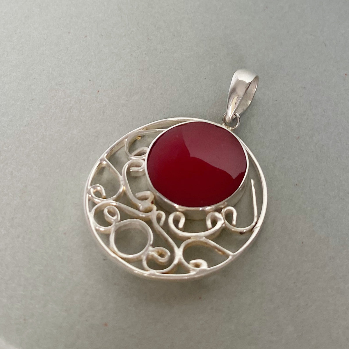 A silver filigree pendant featuring a round, polished deep red gemstone in the center, set against a neutral gray background.