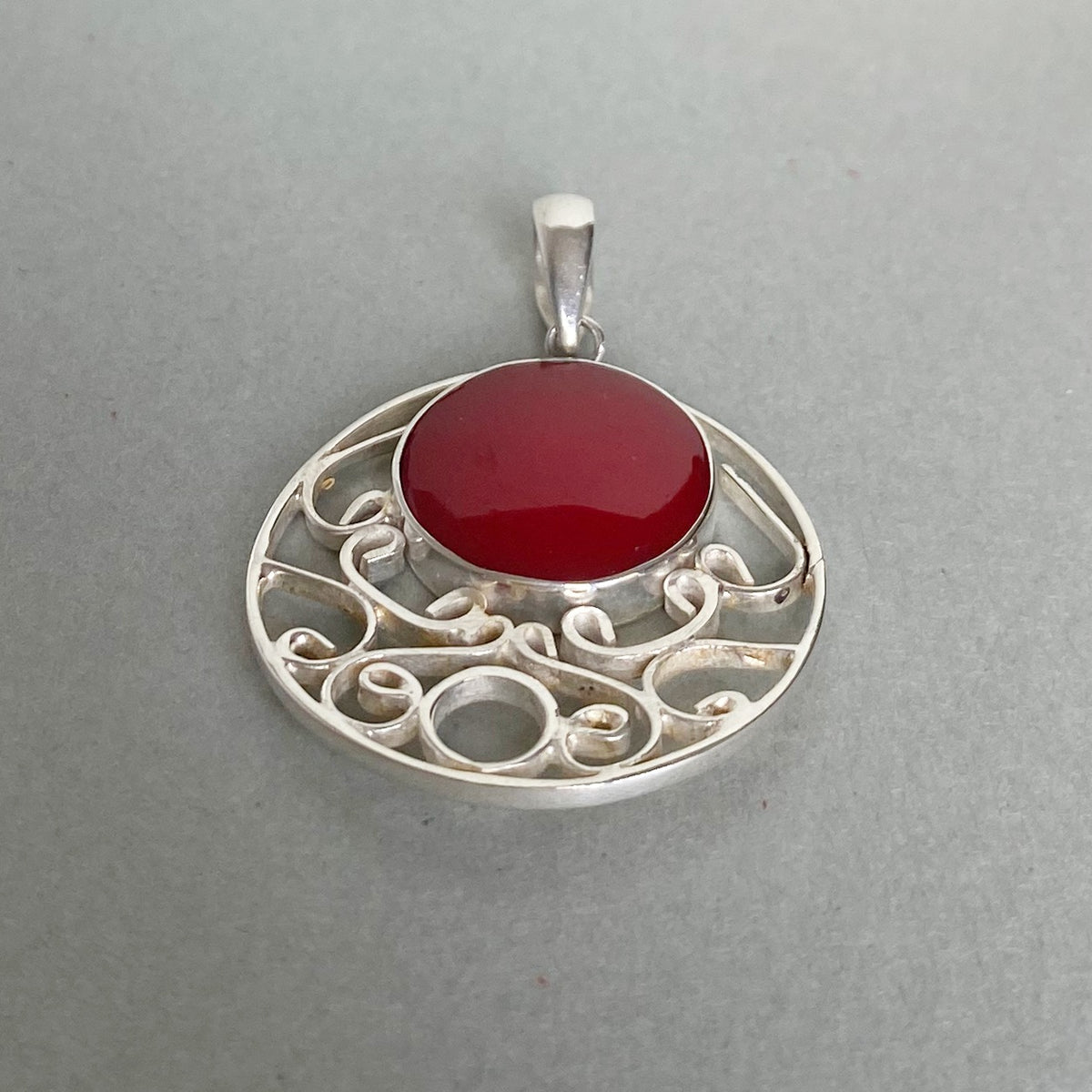 A silver filigree pendant featuring a smooth, oval-shaped deep red gemstone in the center, set against a plain gray background.