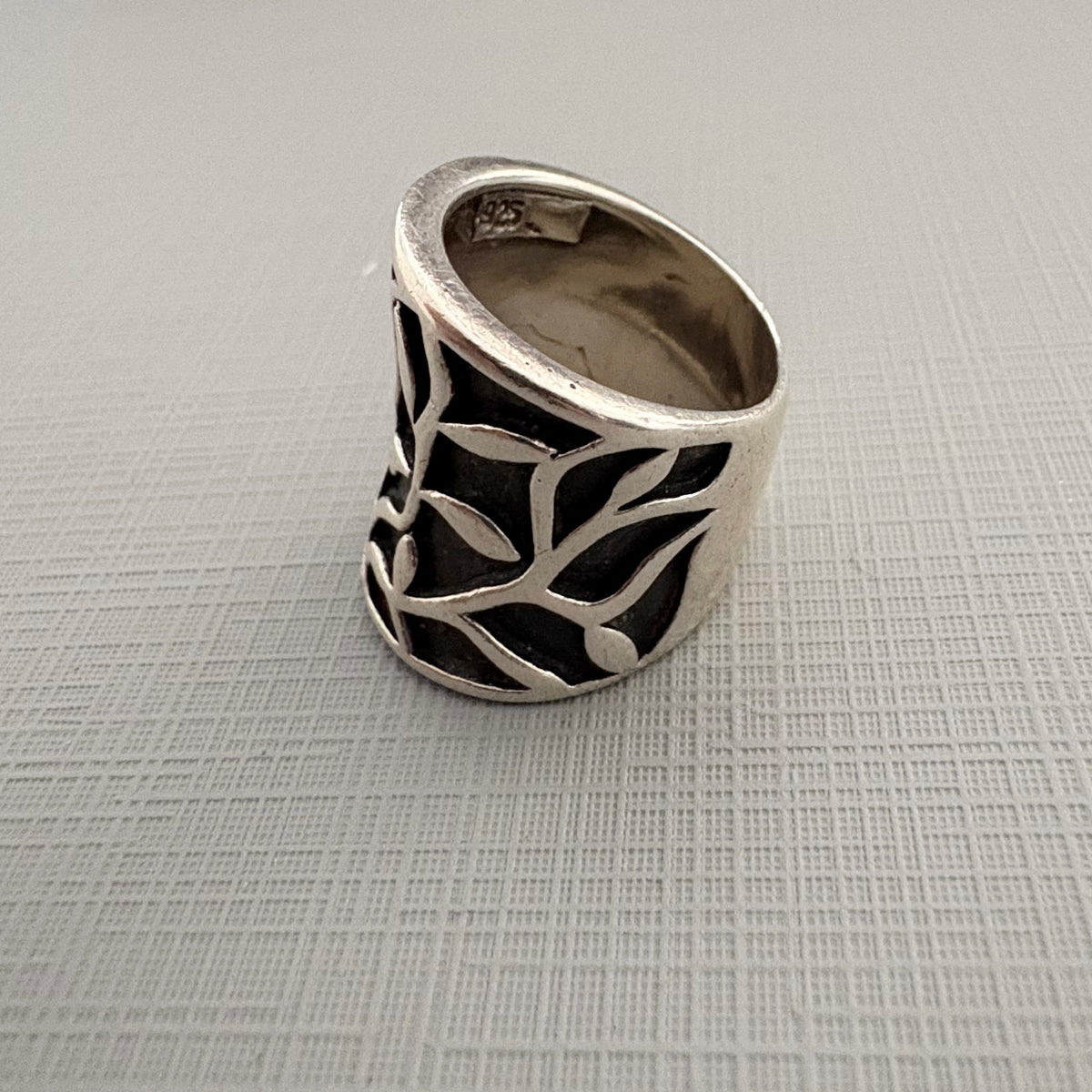 A wide silver ring with a leafy branch filigree cutout design, displayed on a textured light gray surface.