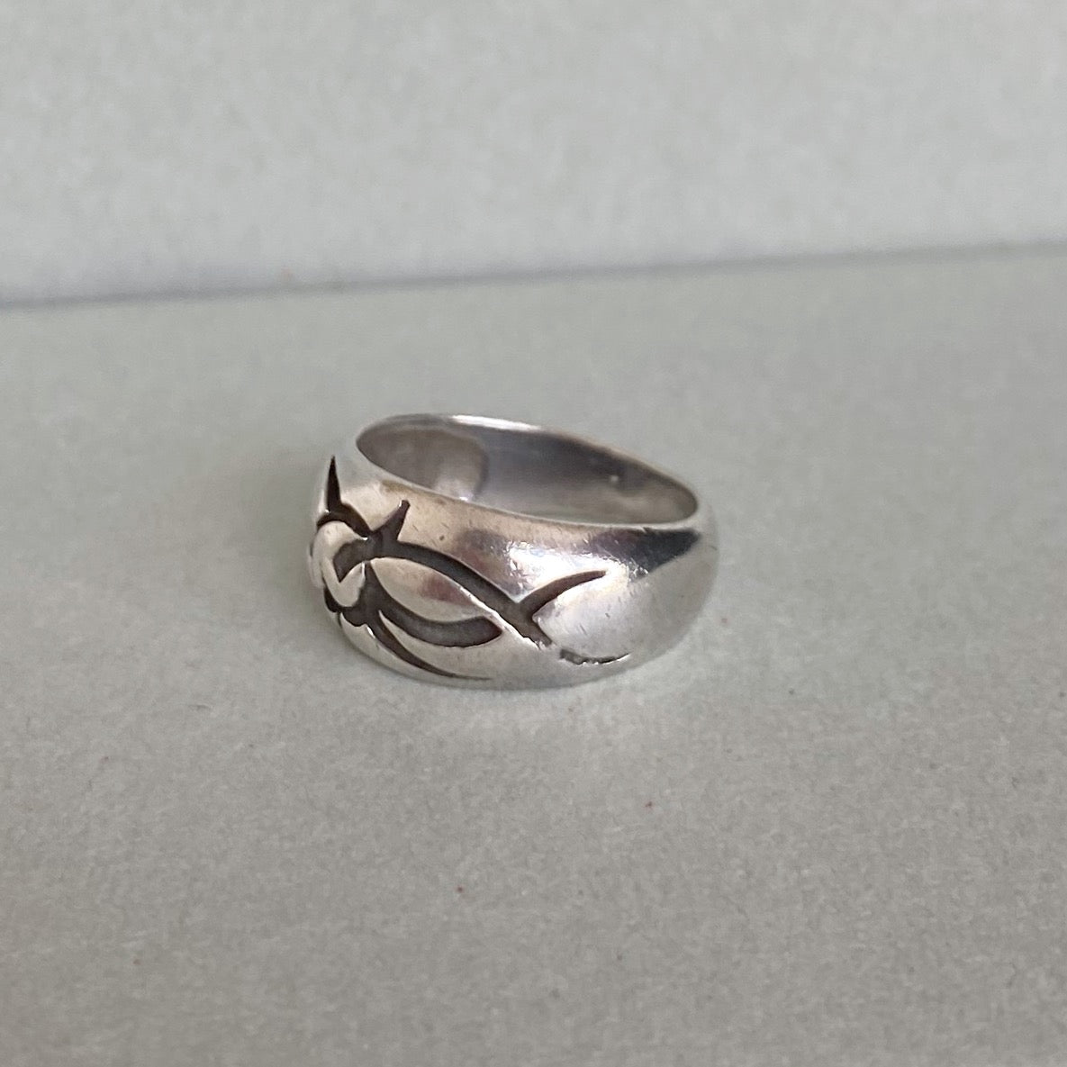 A sterling silver band ring with a stylized cutout tribal or wave-like pattern on the front, resting on a neutral gray surface.