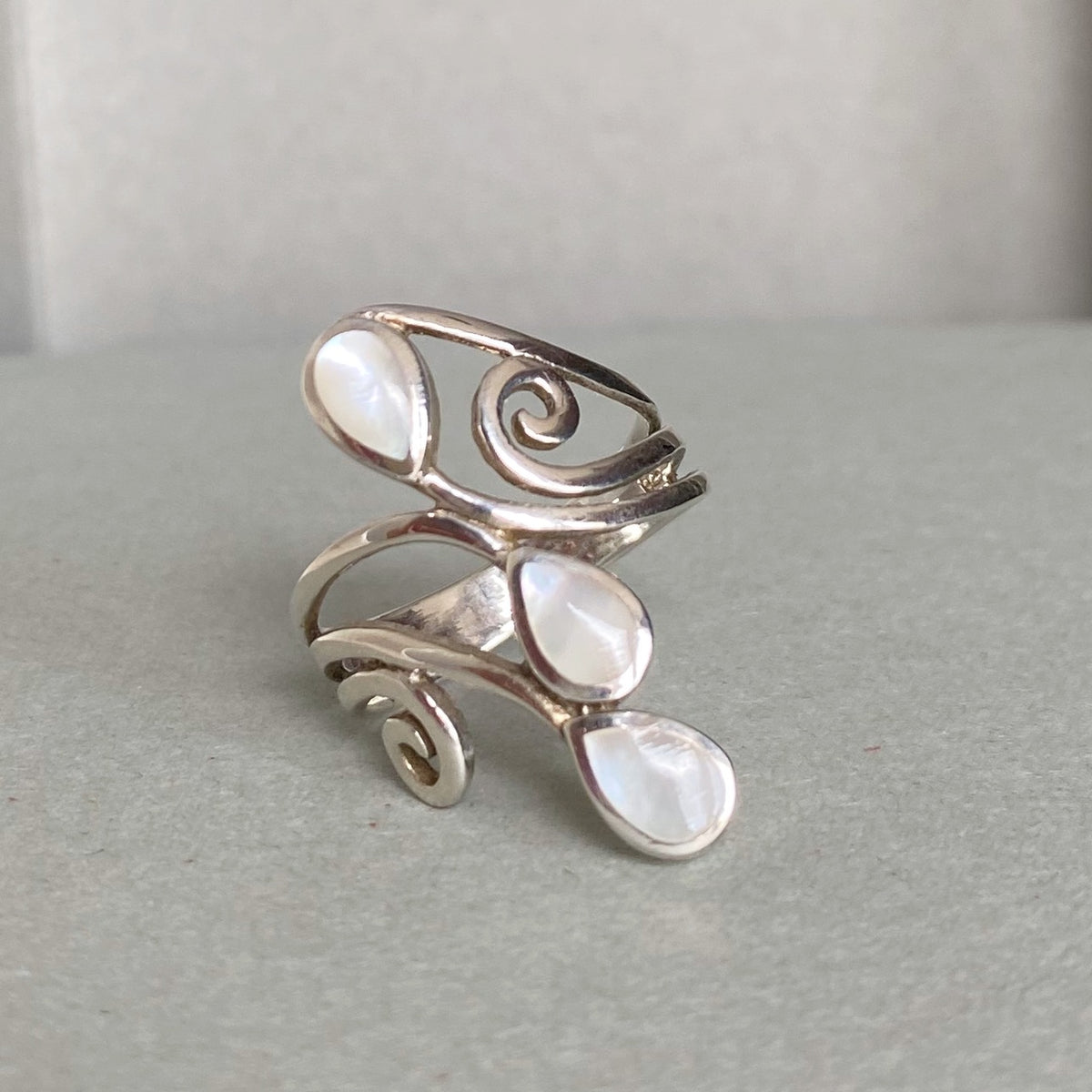 A silver wrap ring featuring three teardrop-shaped white mother-of-pearl inlays, displayed on a neutral gray surface.