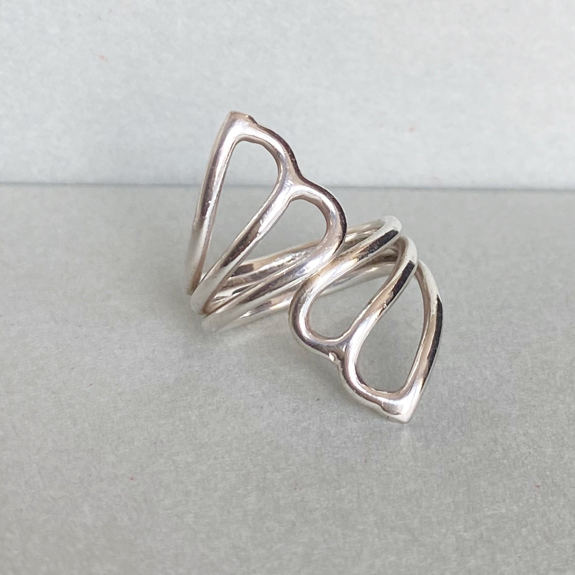 A polished silver openwork ring with a sculptural, flowing loop design, displayed on a neutral gray surface.