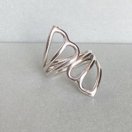 A polished silver openwork ring with a sculptural, flowing loop design, displayed on a neutral gray surface.