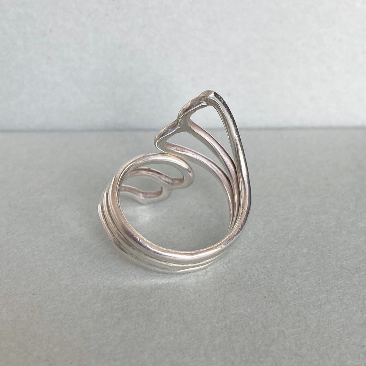 A silver-toned open ring with a stylized, flowing abstract design, photographed on a neutral gray surface against a light gray background.