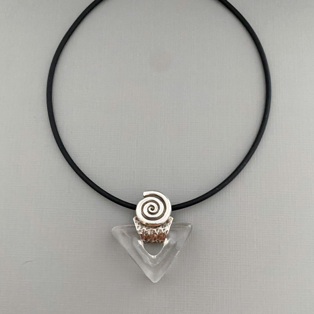 A black cord necklace featuring a clear triangular crystal pendant with a textured silver spiral accent at the top, displayed on a neutral gray background.