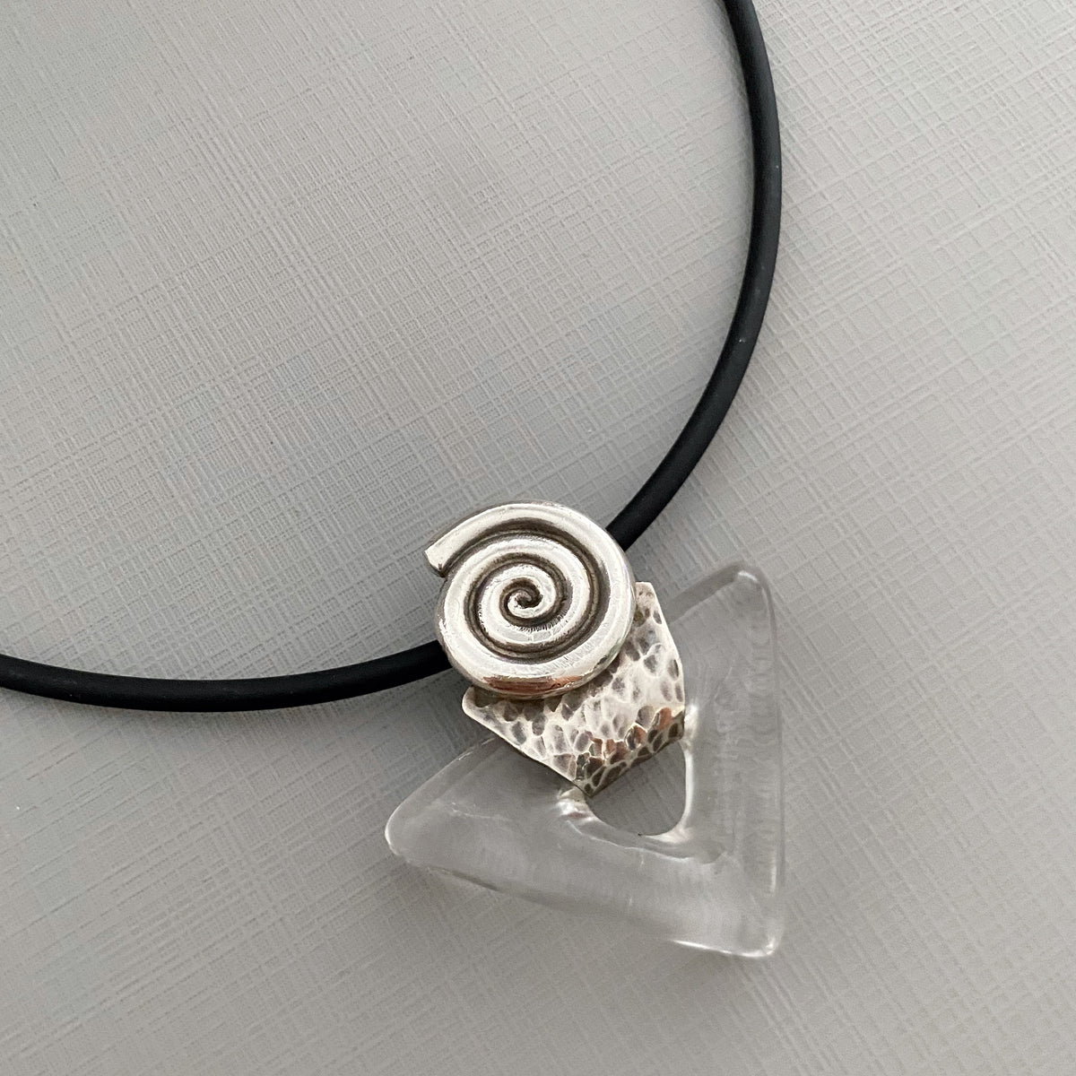A close-up of a necklace with a black cord, a hammered silver spiral charm, and a clear triangular glass bead on a light gray textured background.