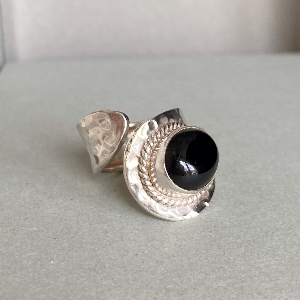 A silver ring featuring a large, polished black onyx cabochon stone in a textured, hammered bezel setting, photographed on a neutral gray surface.