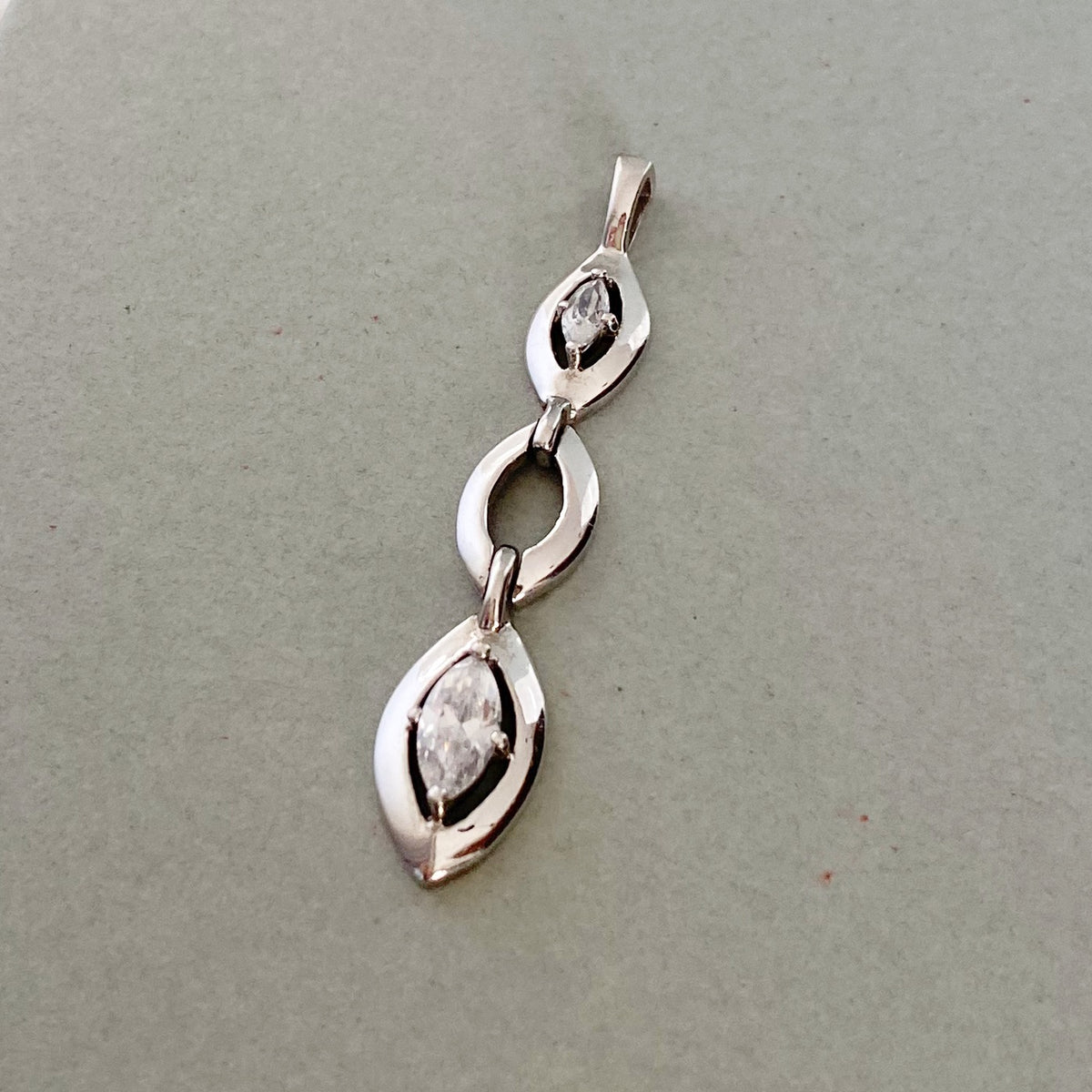A silver pendant featuring three stacked teardrop-shaped links, each holding a small marquise-cut clear gemstone, displayed on a neutral gray background.