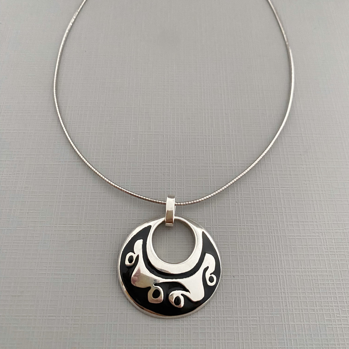 A silver pendant necklace featuring a circular disc with a cutout center and stylized silver swirls on a black background, suspended from a thin silver chain on a textured gray background.