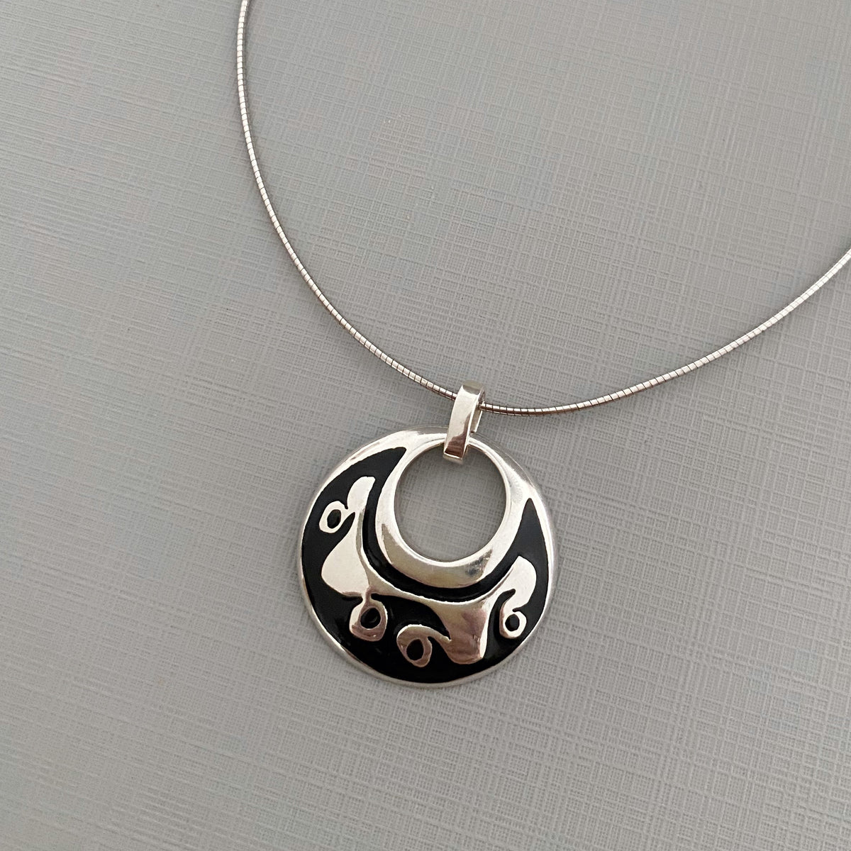 A sterling silver pendant necklace featuring a circular disc design with a central cutout and abstract engraved details, displayed on a textured gray background.