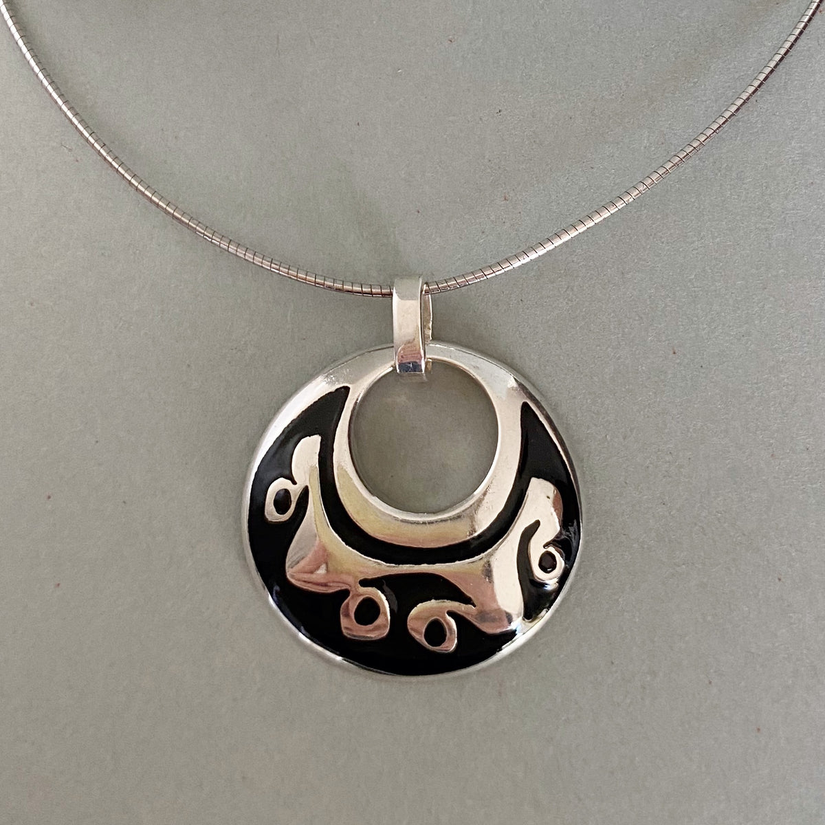 A silver pendant necklace featuring a circular disc design with a cutout crescent moon and abstract wave patterns over a black background, suspended from a silver snake chain on a neutral gray background.