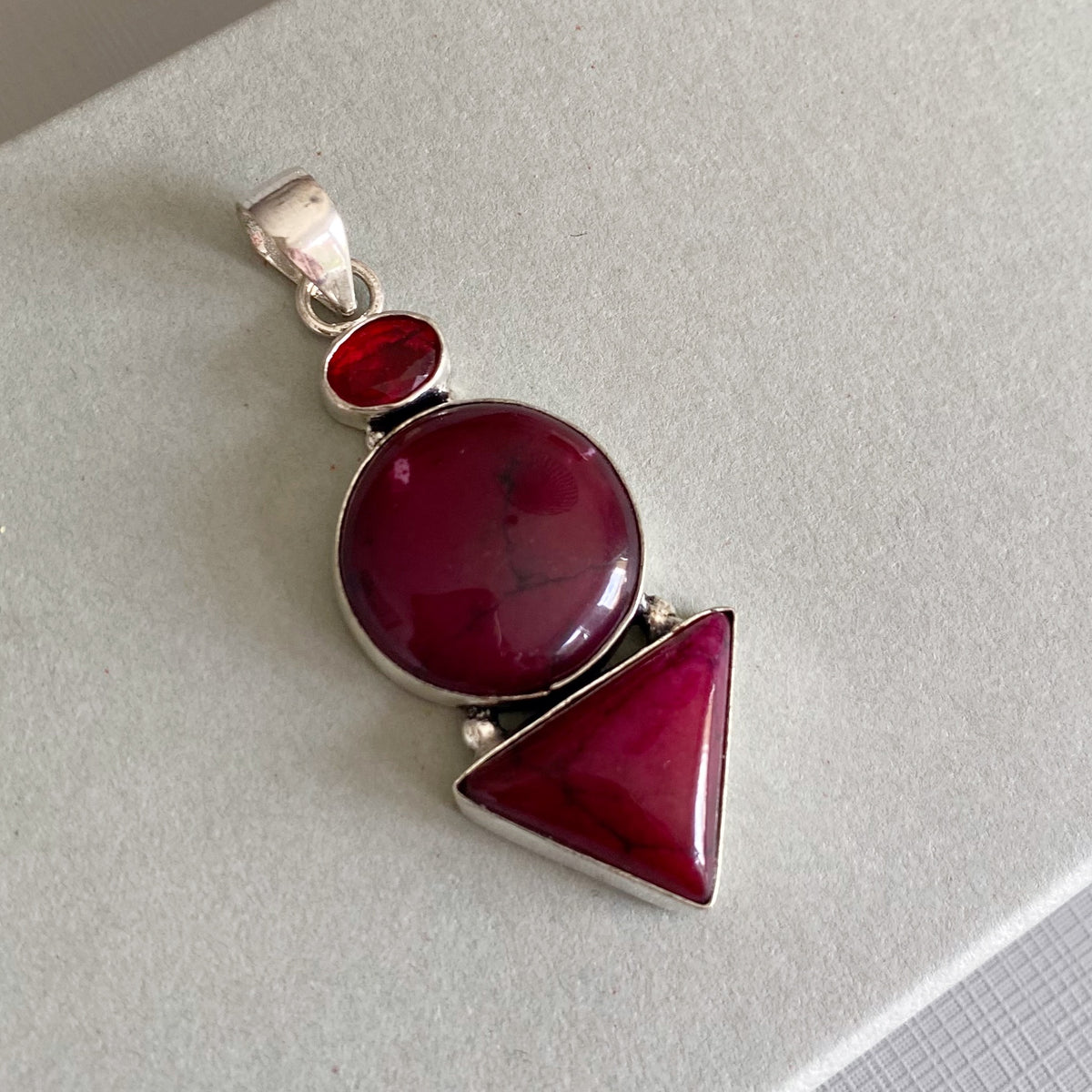 A silver pendant featuring three stacked geometric gemstone shapes in deep magenta-red, resting on a light gray textured surface.
