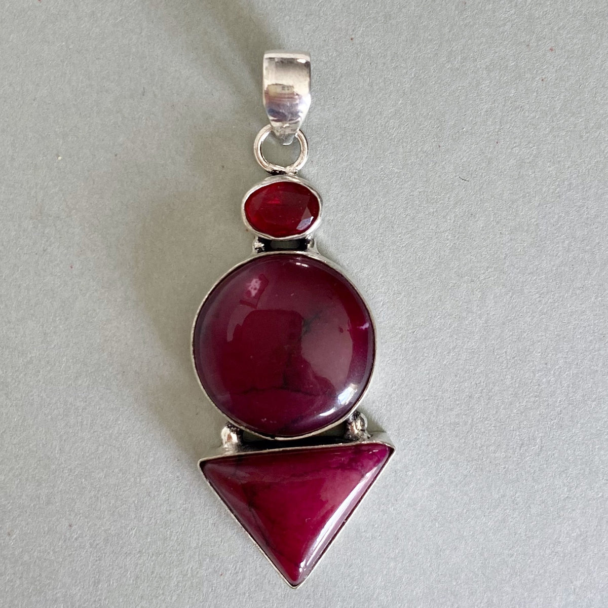 A silver pendant featuring three stacked gemstone shapes: a small oval red stone at the top, a larger oval deep magenta-red stone in the middle, and a pointed triangle in the bottom, set in polished silver on a light gray background.