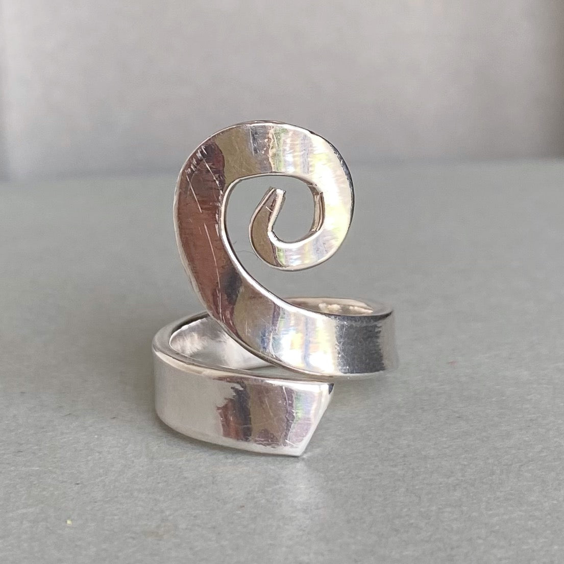 A polished silver ring with a sculptural, open spiral design, displayed on a neutral gray surface.