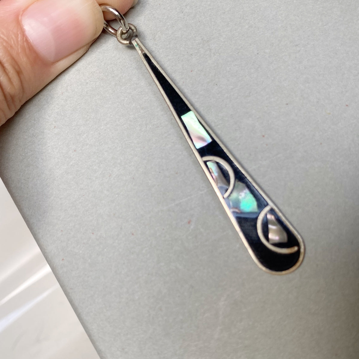 A close-up of a silver teardrop pendant with black inlay and iridescent abalone shell accents, held by a hand against a gray background.