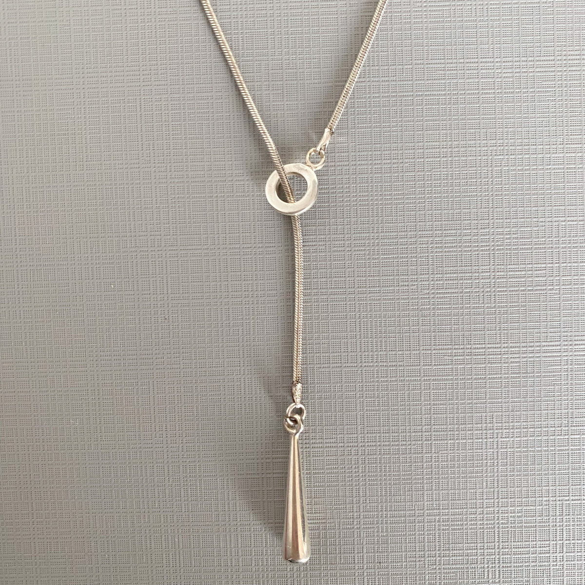 A silver-toned Y-shaped necklace with a flat circular ring at the top and a tapered teardrop pendant hanging below, displayed on a textured gray background.