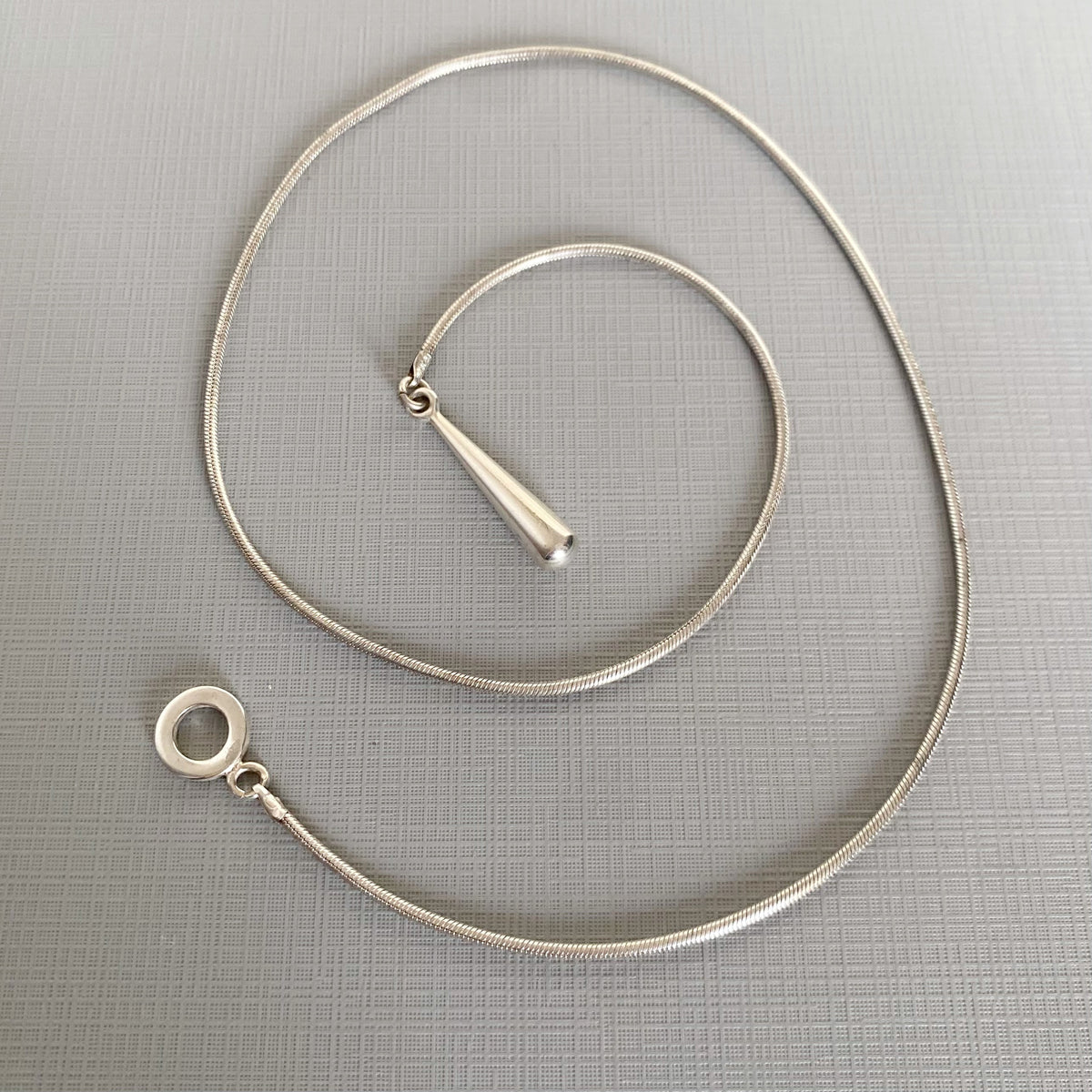 A silver-toned snake chain necklace with a teardrop-shaped pendant and a circular ring clasp, displayed on a textured gray background.