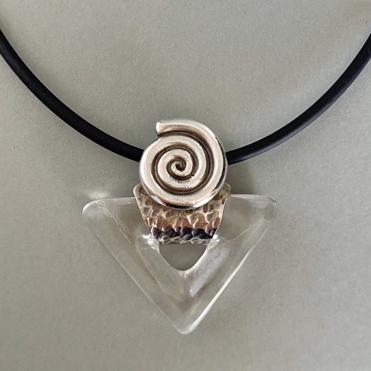A handmade pendant necklace featuring a spiral shell charm in a silver-toned setting with a hammered rose-gold textured base, suspended on a black cord over a clear triangular glass holder against a neutral gray background.